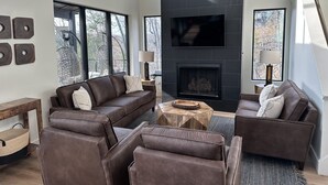 Ein modernes Wohnzimmer mit Kamin, Ledersofas und großen Fenstern mit Blick auf Bäume.