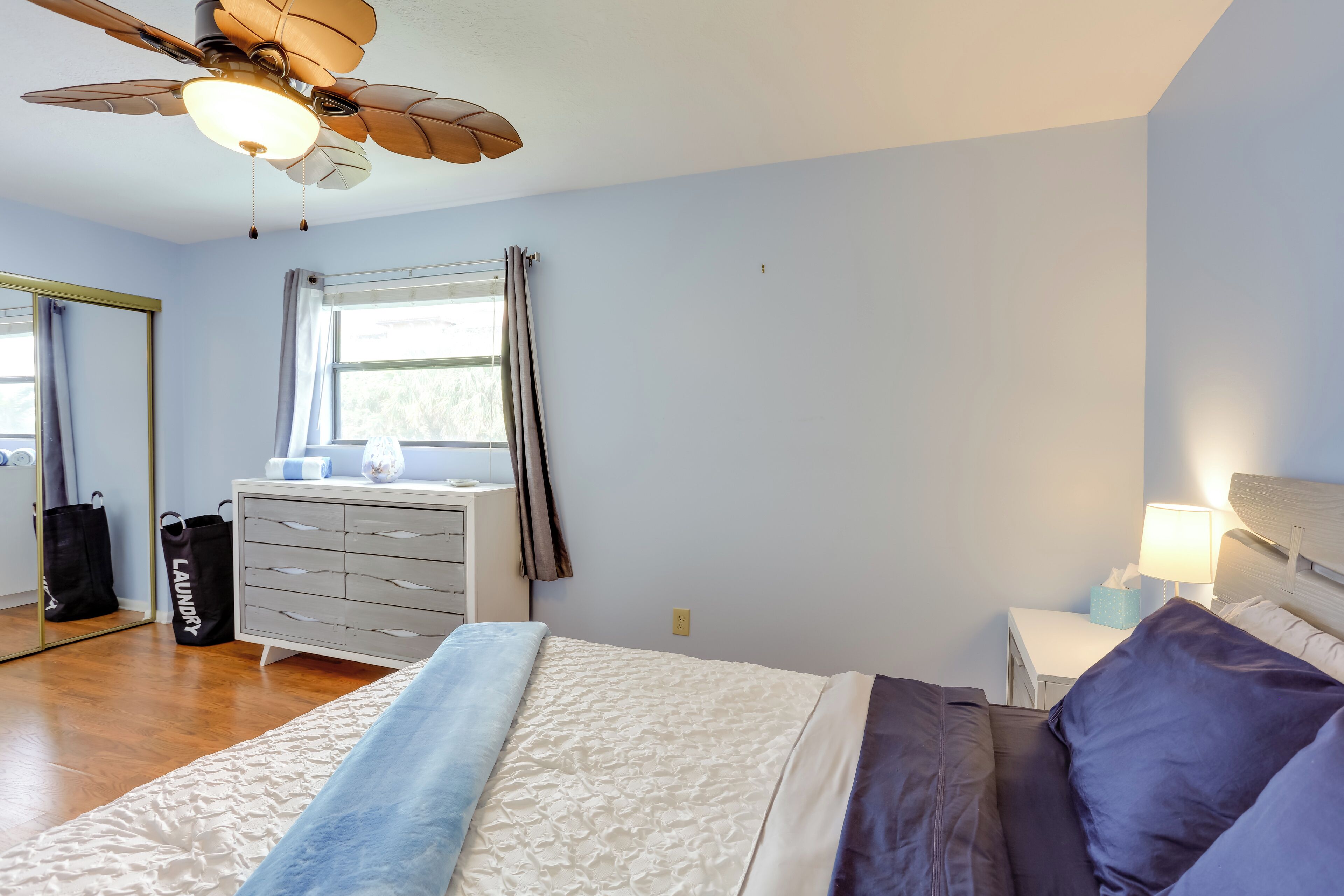 A bedroom with a bed, a dresser, a nightstand, a ceiling fan, and a window with curtains.