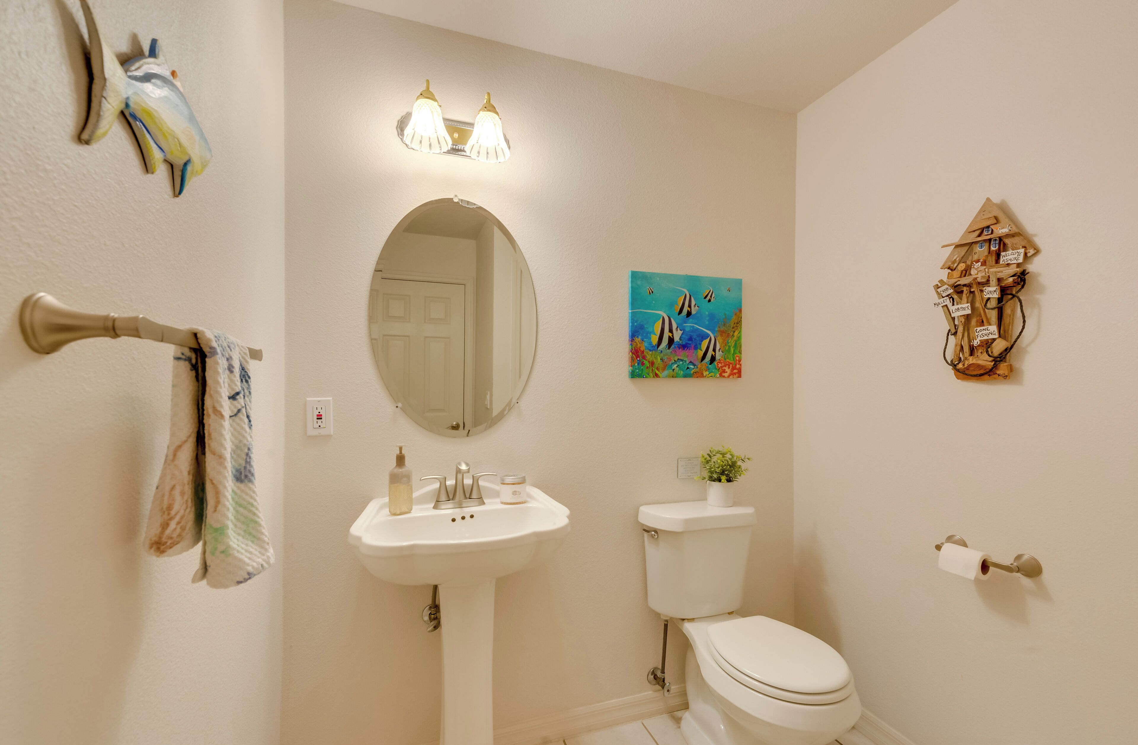 A bathroom with a sink, toilet, and a mirror. There is a towel rack with a towel, a painting on the wall, and a decorative item above the toilet.