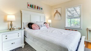 A neatly arranged bedroom with a white bed, two bedside tables with lamps, a decorative wall piece, and a window with blinds. - Pensacola Home w/ Covered Patio: 1 Mi to Downtown! (Pensacola)