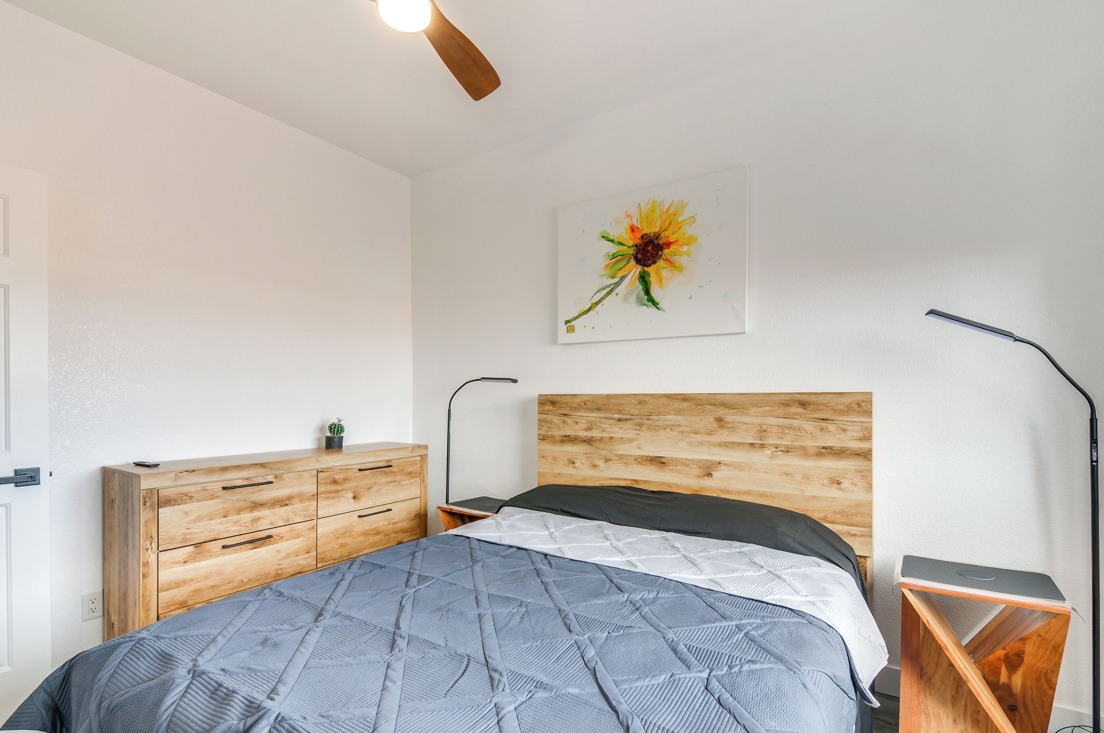 A bedroom with a wooden headboard, a bed with a quilt, a nightstand, a desk, a lamp, and a painting of a sunflower.