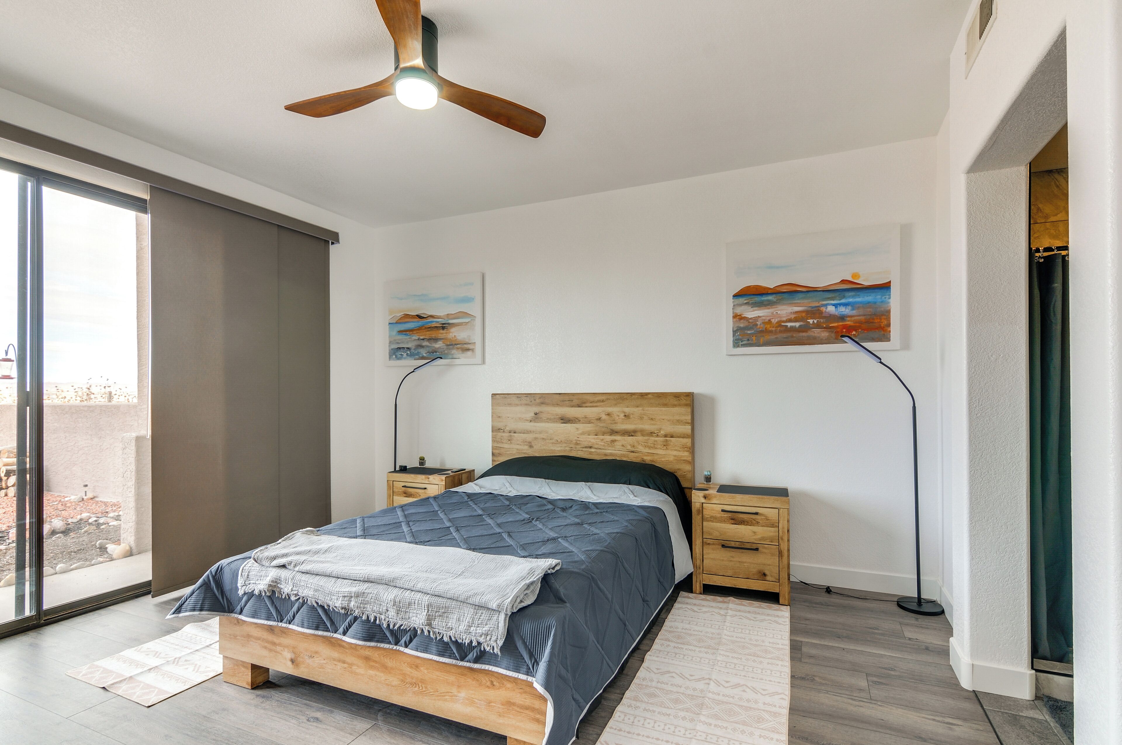 A bedroom with a bed, bedside tables, a ceiling fan, artwork on the walls, and a view through a sliding door.