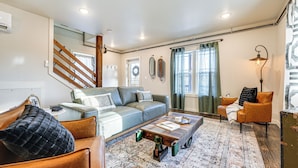 Un salon moderne avec un canapé, une table basse et un fauteuil. On y trouve un escalier avec des rampes en bois et une vue sur l’extérieur par une fenêtre ornée de rideaux.