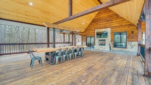 Eine geräumige Holzterrasse mit einem langen Tisch und Stühlen, einem Steinkamin und großen Fenstern mit Blick auf einen Wald.