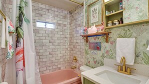 A bathroom with a brick wall, a pink bathtub, a toilet, a sink with a gold faucet, and a shower curtain with floral patterns. - Pet-friendly Brownwood Home: Walk to Lake! (Brownwood)