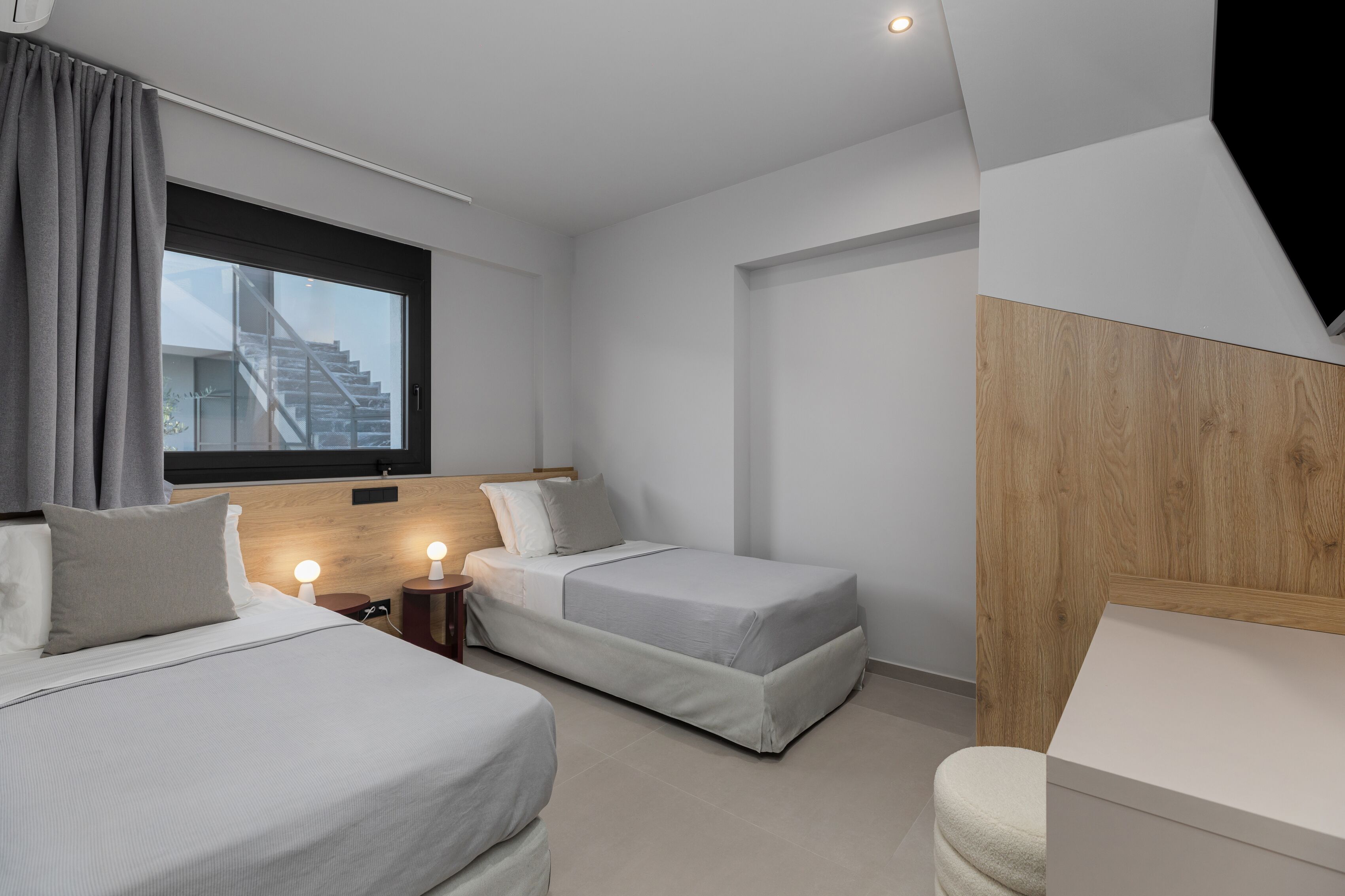 A modern hotel room with two beds, a wooden headboard, a large window with a view of a staircase, and a small bedside table with a lamp.