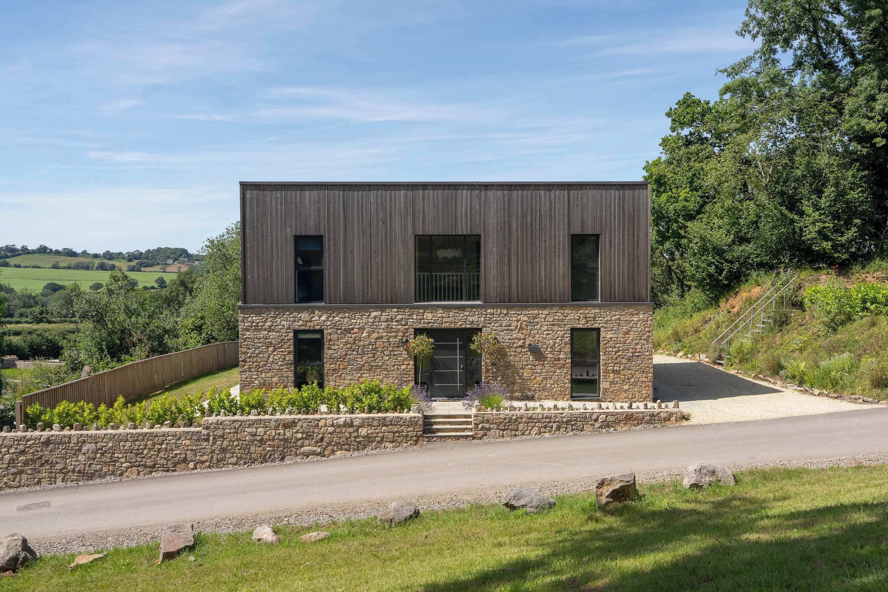 Una casa moderna de dos pisos con fachada de piedra, amplias ventanas y un camino de entrada a un acceso con puerta.