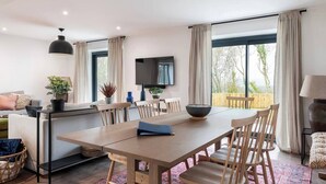 Un salon moderne comprenant un coin repas, un téléviseur fixé au mur et de grandes fenêtres avec des rideaux.