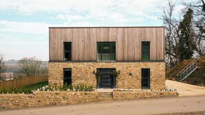 Une maison moderne à deux étages, dotée d’une façade en pierre et d’étages supérieurs en bois, avec une entrée principale accessible par des marches et un petit jardin.