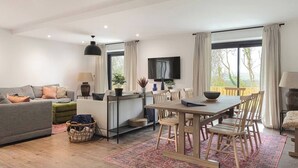 Un salon moderne comprenant un canapé, une table à manger avec des chaises, un téléviseur fixé au mur et une vue sur l’extérieur grâce à de grandes fenêtres.