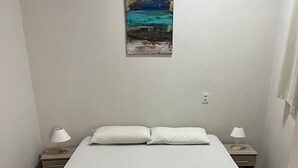 Ein Einzelbett mit weißer Matratze und braunem gesteppten Bettlaken, begleitet von zwei Nachttischen mit Lampen, in einem Zimmer mit schlichten weißen Wänden und einem Fliesenboden.
