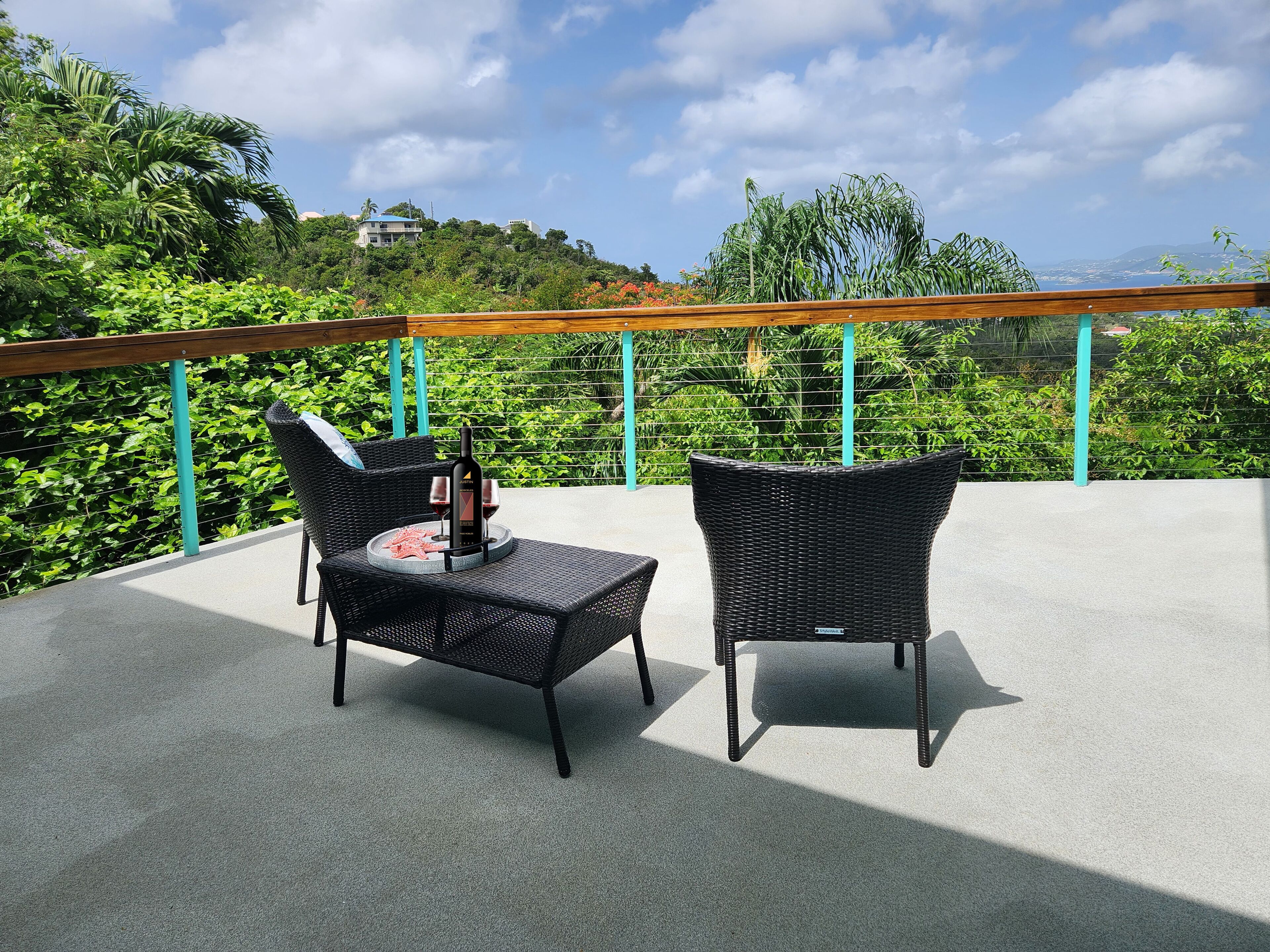 Een balkon met twee rieten stoelen, een tafel met een fles wijn en glazen, en een uitzicht op weelderige begroeiing en gebouwen op de achtergrond.
