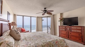A hotel room with a large bed, a ceiling fan, a TV on a wooden dresser, and a view of the ocean through the sliding glass door.