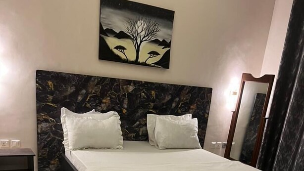 A bedroom with a bed, two pillows, a nightstand, a bench, a small table, a mirror, and a painting on the wall. - Anastera Home Vila (Kigali)