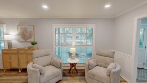 A room with two upholstered chairs, a small round table with a lamp, a wooden cabinet, and a large window with blinds. - A Lifelong Dream - 3 bedroom, 2 bath with private swimming pool (Jekyll Island)
