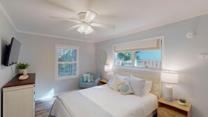 A bedroom with a bed, bedside tables, a ceiling fan, a TV, a window with blinds, and a view of the outdoors. - A Lifelong Dream - 3 bedroom, 2 bath with private swimming pool (Jekyll Island)
