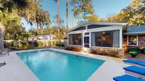 A swimming pool with a covered patio area, surrounded by trees and a brick house. - A Lifelong Dream - 3 bedroom, 2 bath with private swimming pool (Jekyll Island)