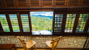 A room with a stone wall, two wooden chairs, and a view of a mountainous landscape through large windows. - Reggae Legends Estate (Kingston)