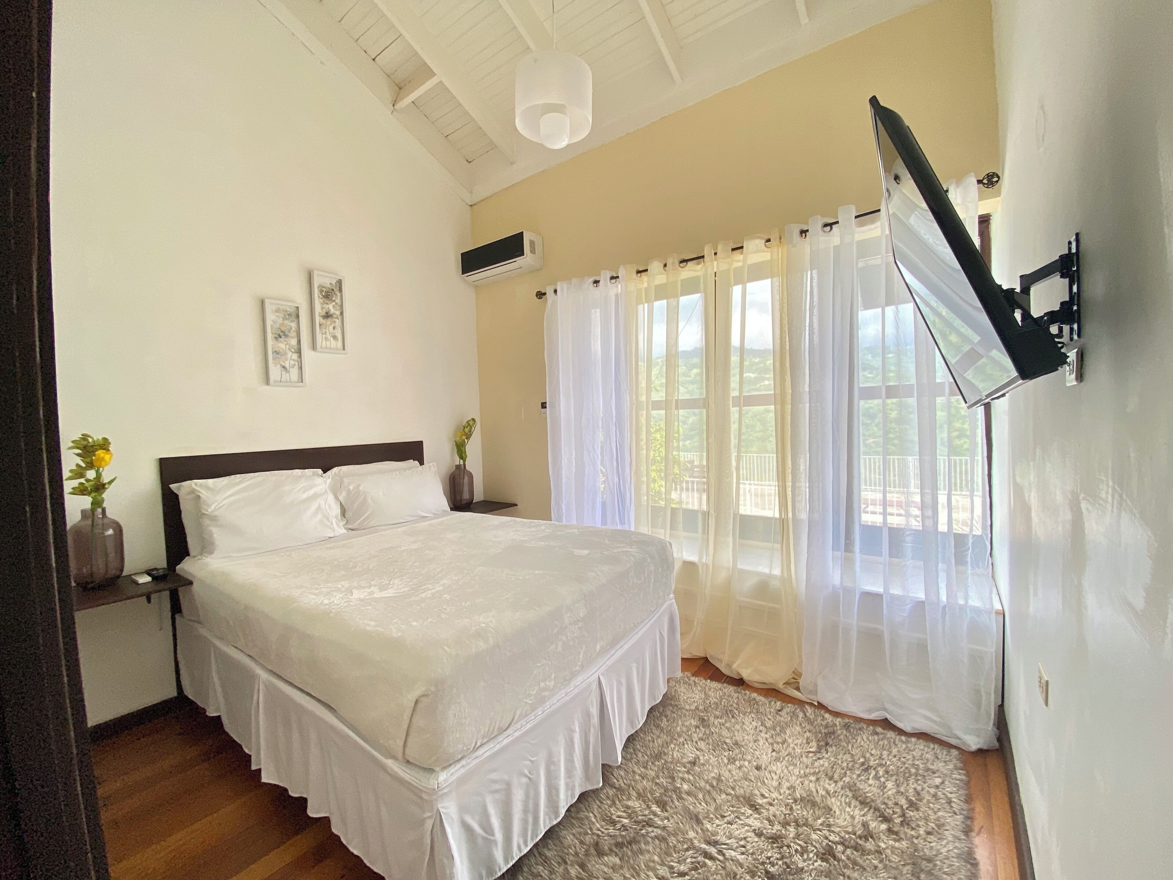 A bedroom with a bed, a nightstand, a wall-mounted TV, and a window with sheer curtains.