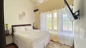 A bedroom with a bed, a nightstand, a wall-mounted TV, and a window with sheer curtains.