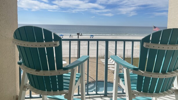 A balcony with two green Adirondack chairs facing a beach with an American flag. - Surf Side Shores 1206  New Sofa   3 bd / 3 bth gulf-front corner unit (Gulf Shores)