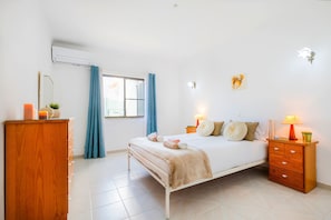 Un dormitorio con cama, mesitas de noche, cómoda de madera, ventana con cortinas e iluminación en la pared.