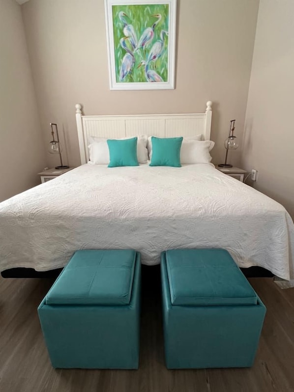 Un dormitorio con cama blanca, dos almohadas color turquesa, cabecero blanco, mesita de noche con lámpara y un cuadro en la pared.