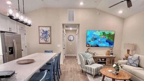 Ein modernes Wohnzimmer mit Sofa, Couchtisch und Fernseher. Es gibt einen Essbereich mit Stühlen und einem Tisch. Der Raum hat Holzböden und einen Deckenventilator.
