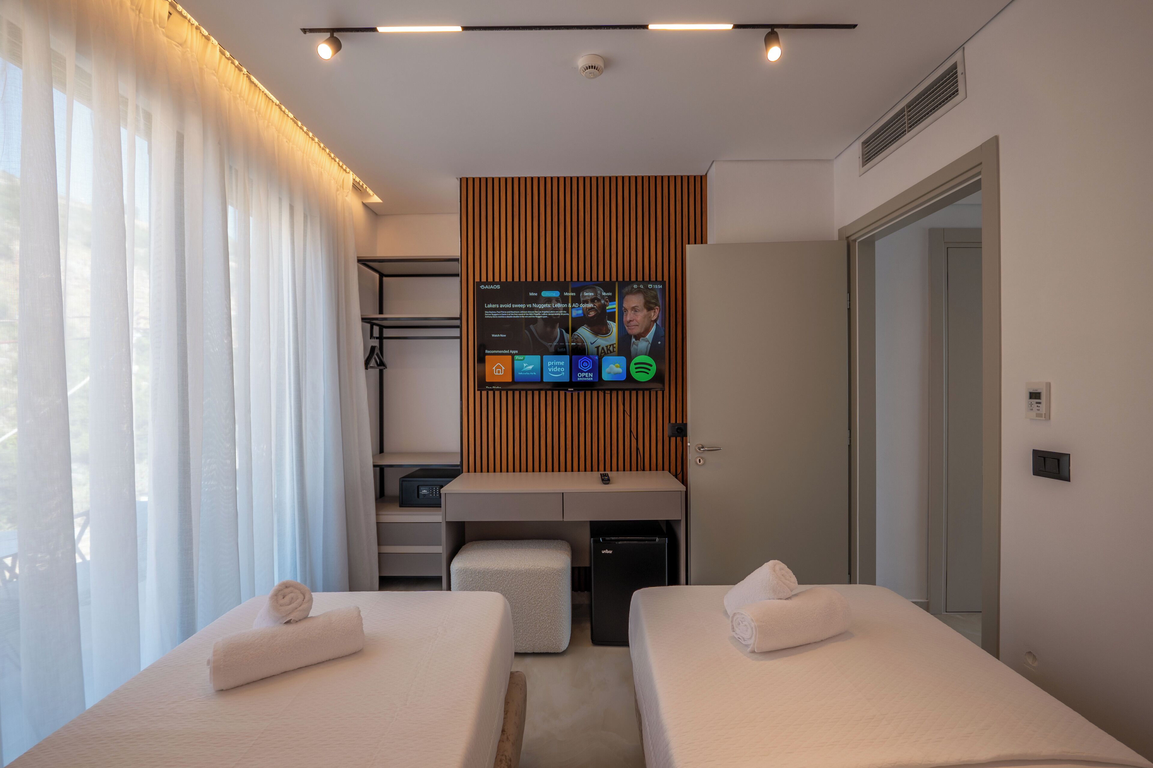 A hotel room with two beds, a desk, a television, and a small kitchenette.