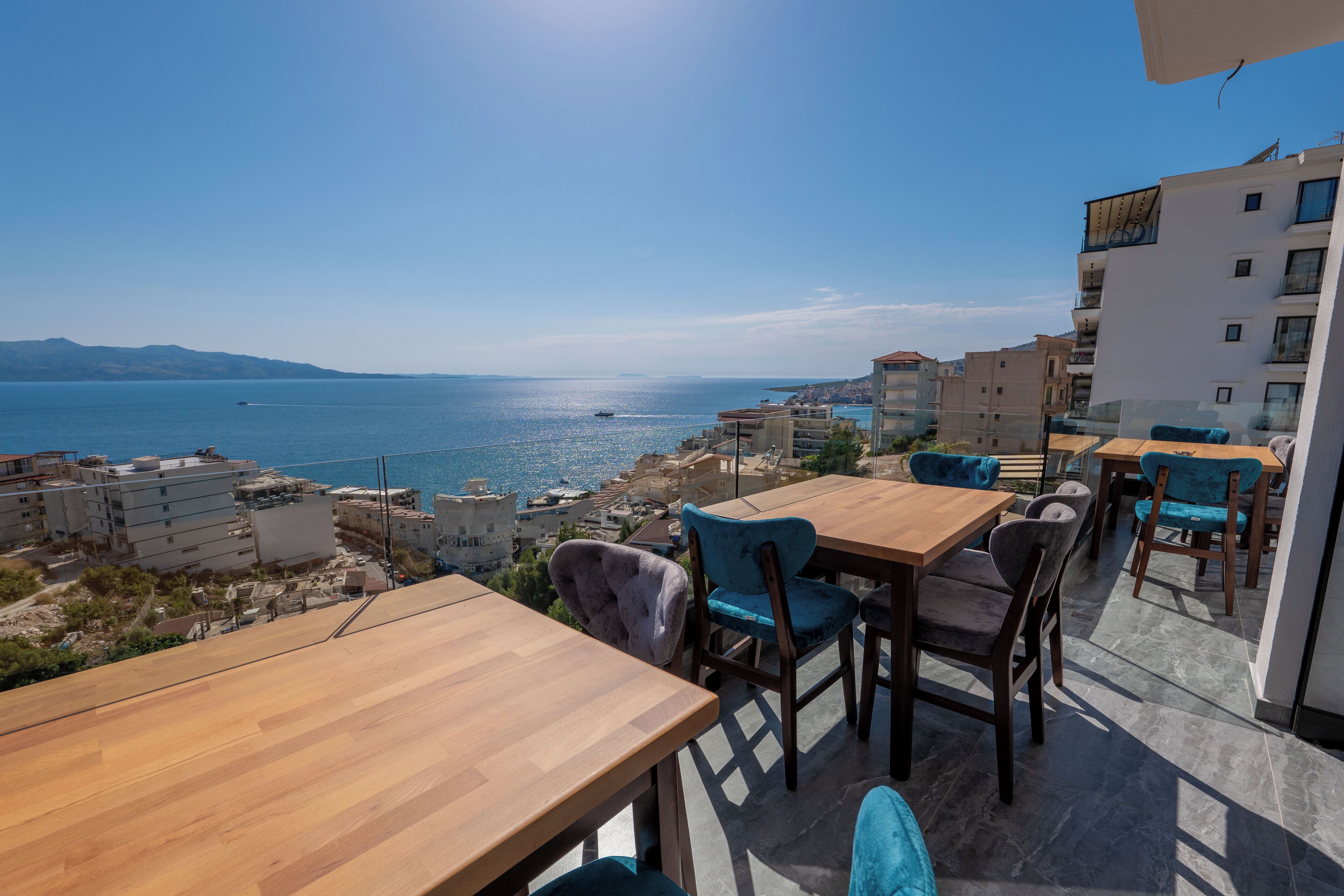 Eine Terrasse mit Holztischen und -stühlen, die Blick auf die Küste mit Gebäuden und dem Meer bietet.