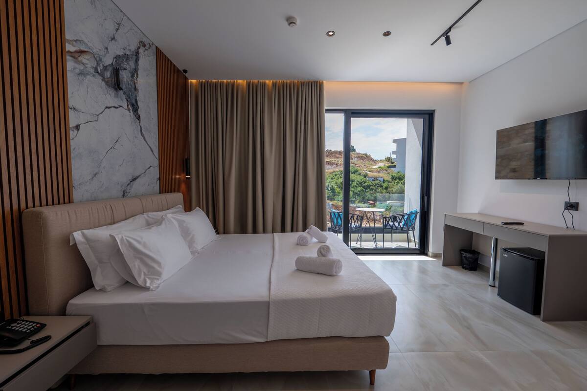 A modern hotel room with a large bed, a desk, a flat-screen TV, and a view of the outdoors.