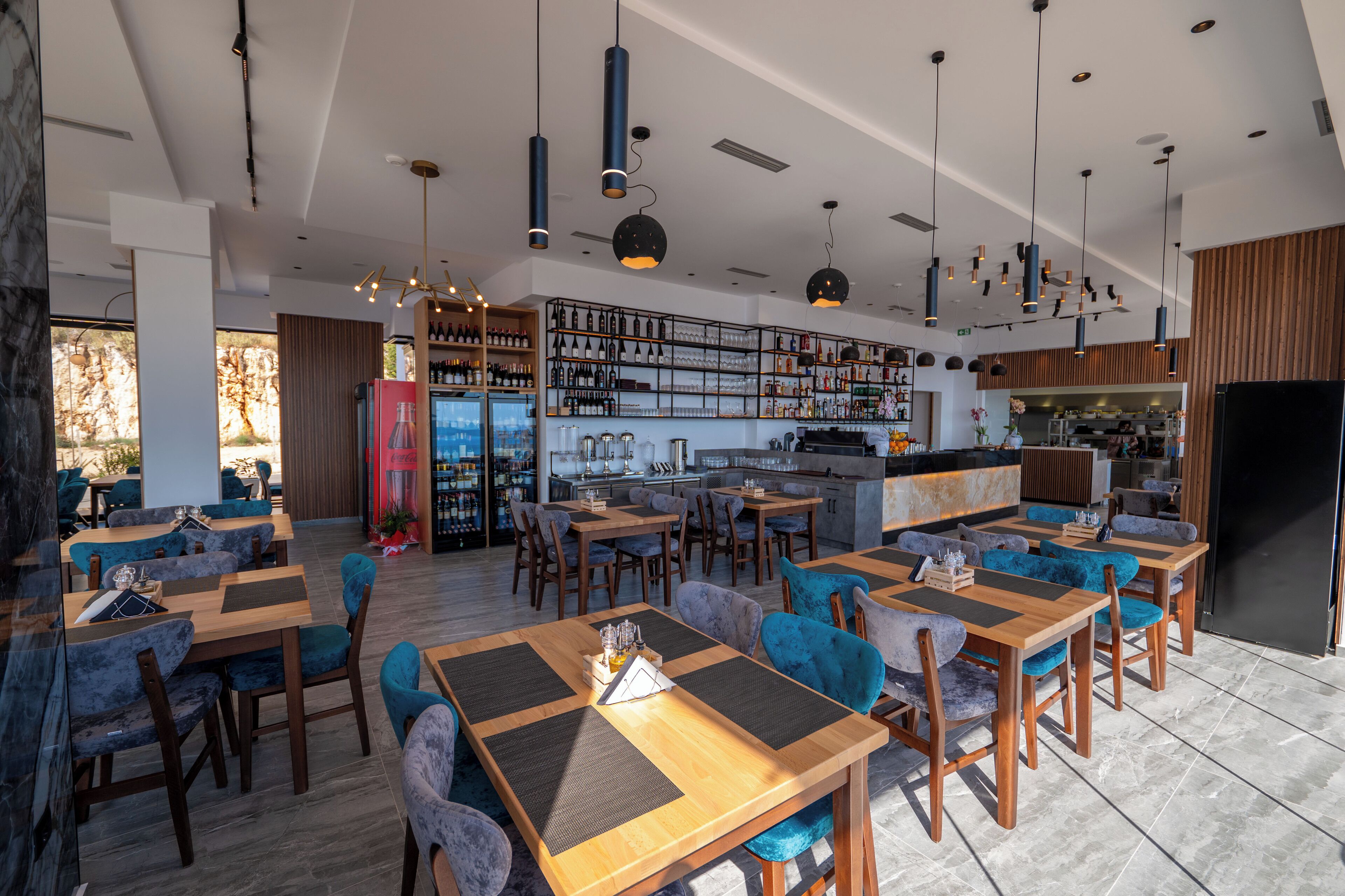 A modern restaurant with wooden tables and blue upholstered chairs, a bar area with shelves of bottles, and a kitchen in the background.