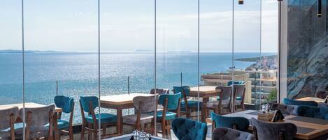 A modern restaurant with large windows offering a view of the sea. Tables and chairs are arranged for dining.