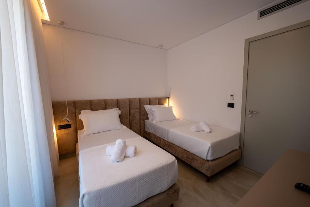 Habitación de hotel con dos camas individuales, una mesita de noche, una puerta y una lámpara fijada a la pared.