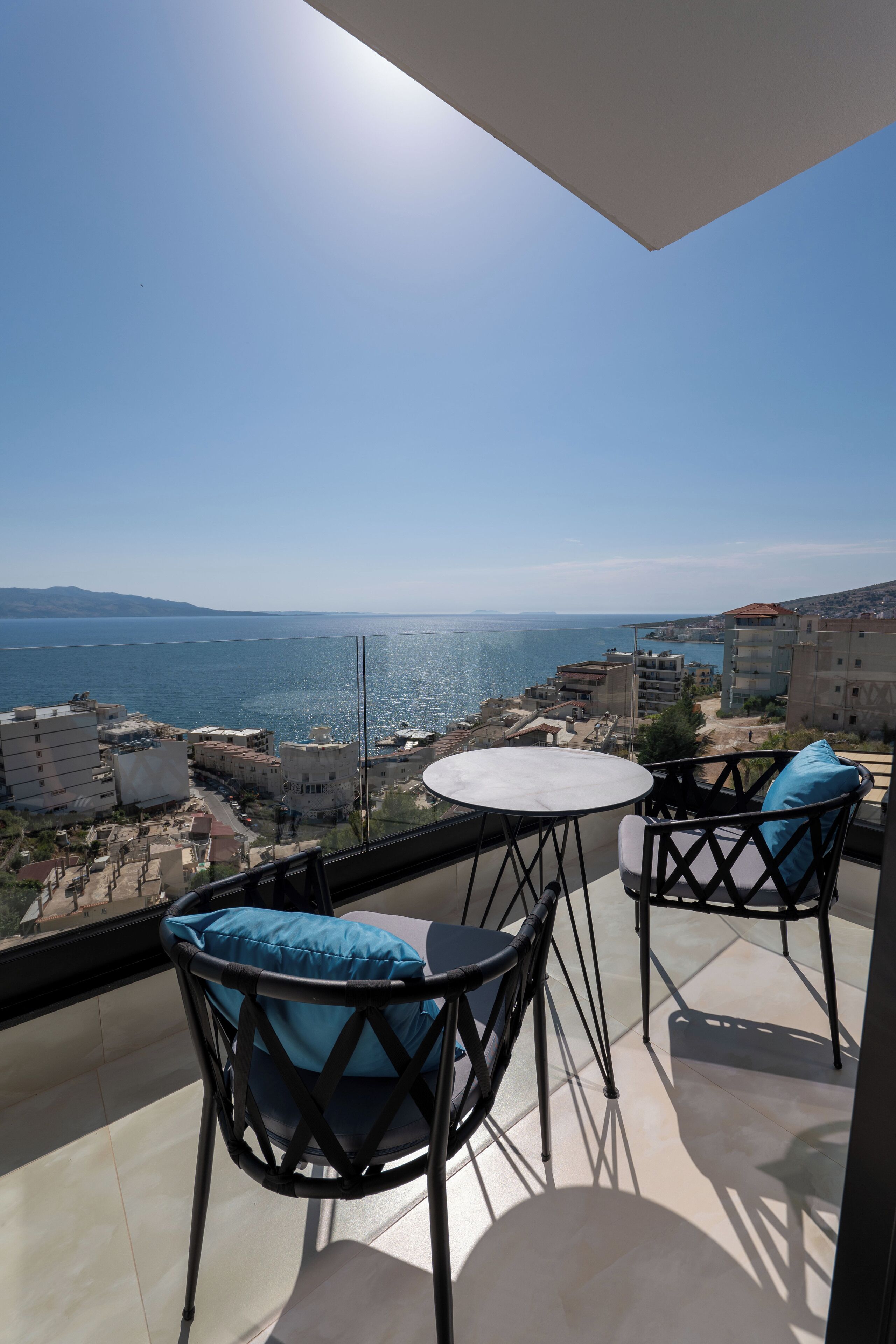 A balcony with two chairs and a table, overlooking a coastal cityscape with buildings and a body of water.