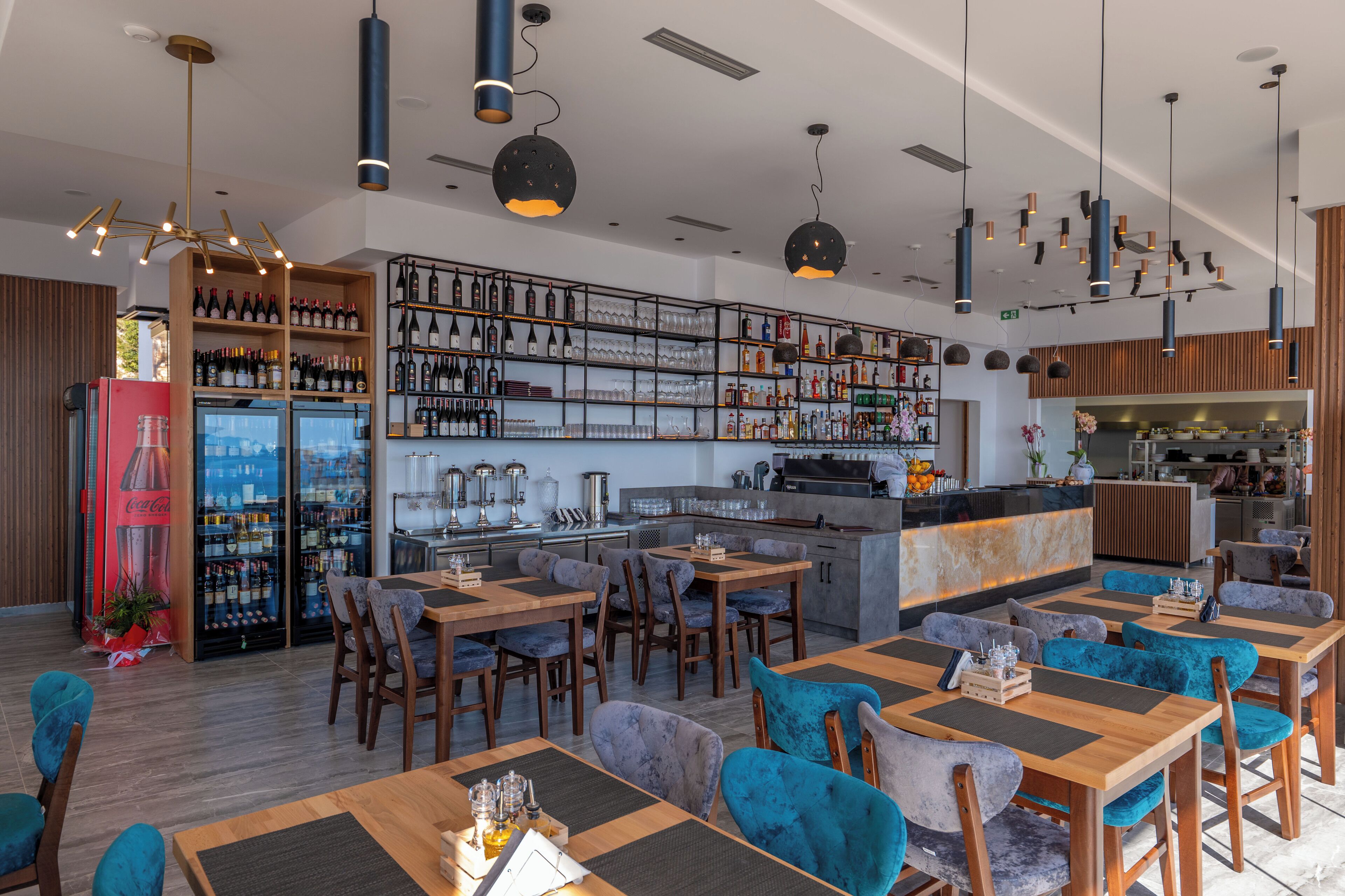 Ein modernes Restaurant mit Holztischen und blauen Stühlen, eine Bar mit Flaschenregalen und eine Küche im Hintergrund.