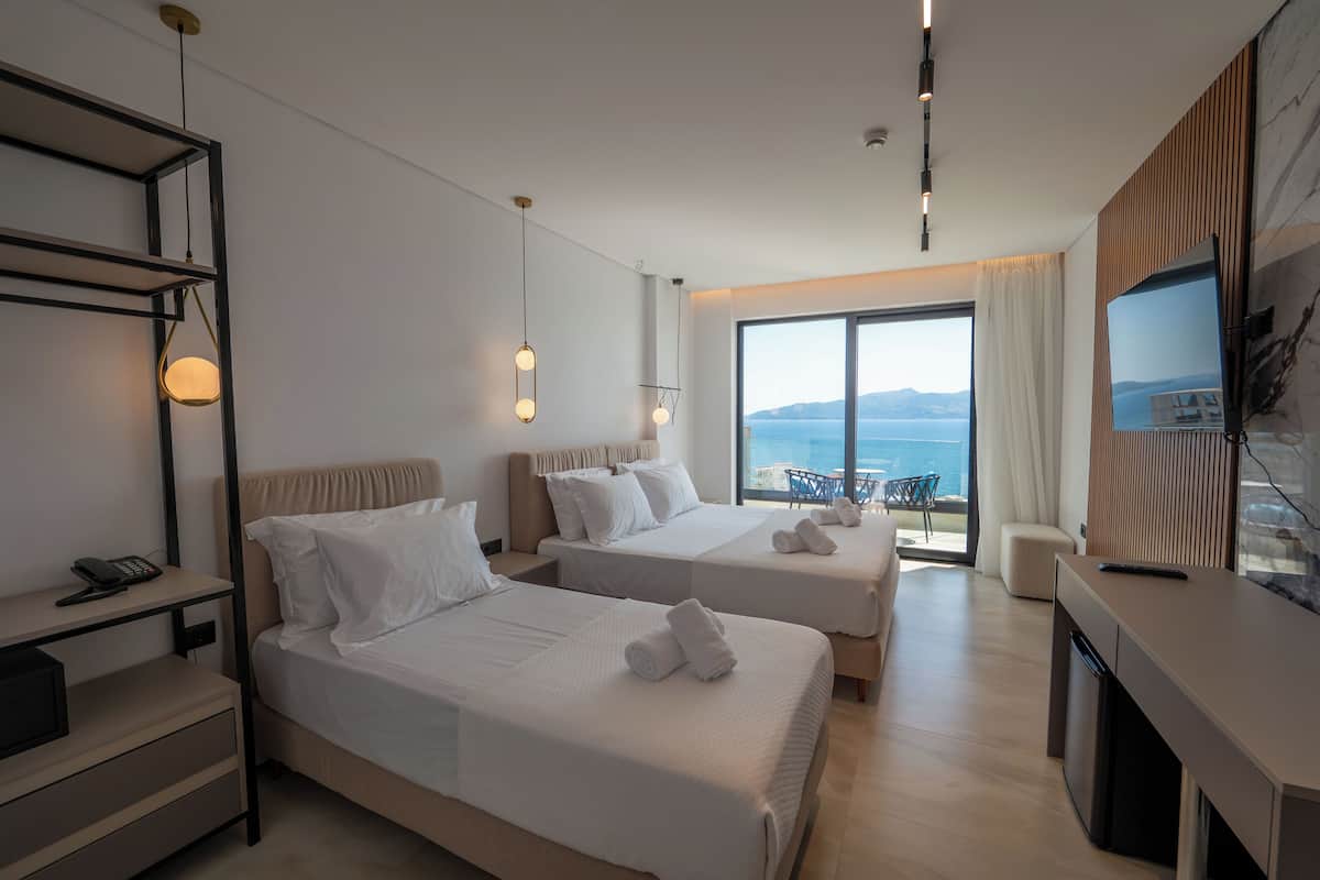 A modern hotel room with two beds, a desk, a TV, and a balcony with a view of the sea.