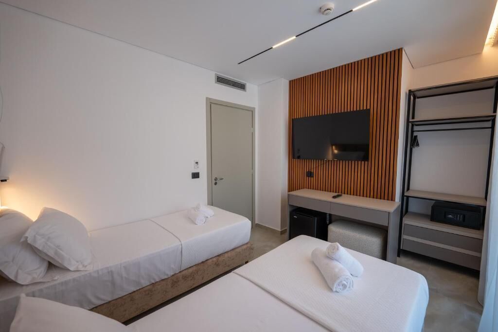 A hotel room with two single beds, a TV mounted on a wooden panel wall, a desk with a chair, and a wardrobe.