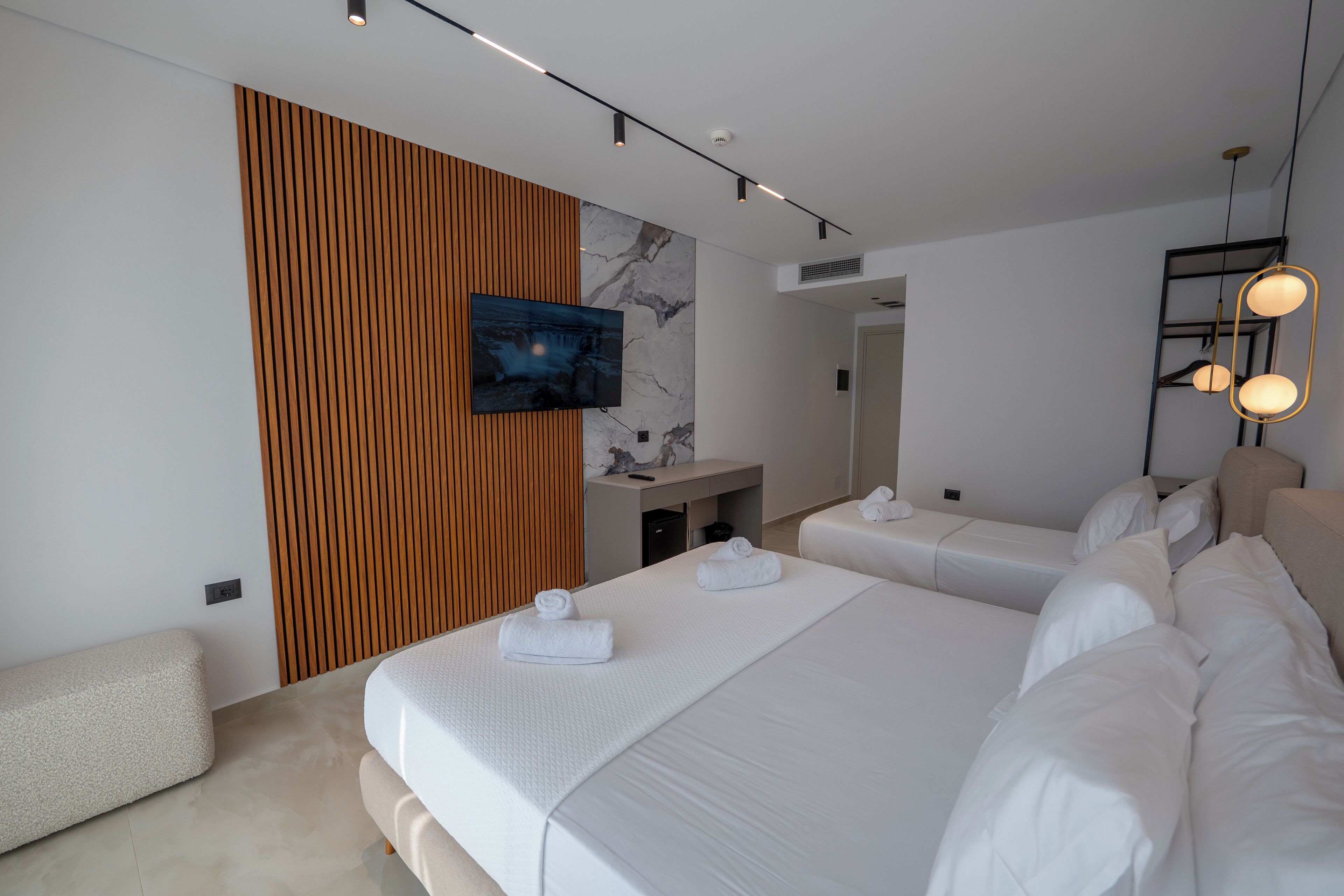 A modern hotel room with two beds, a TV mounted on a wooden panel, a small desk, and a mirror.