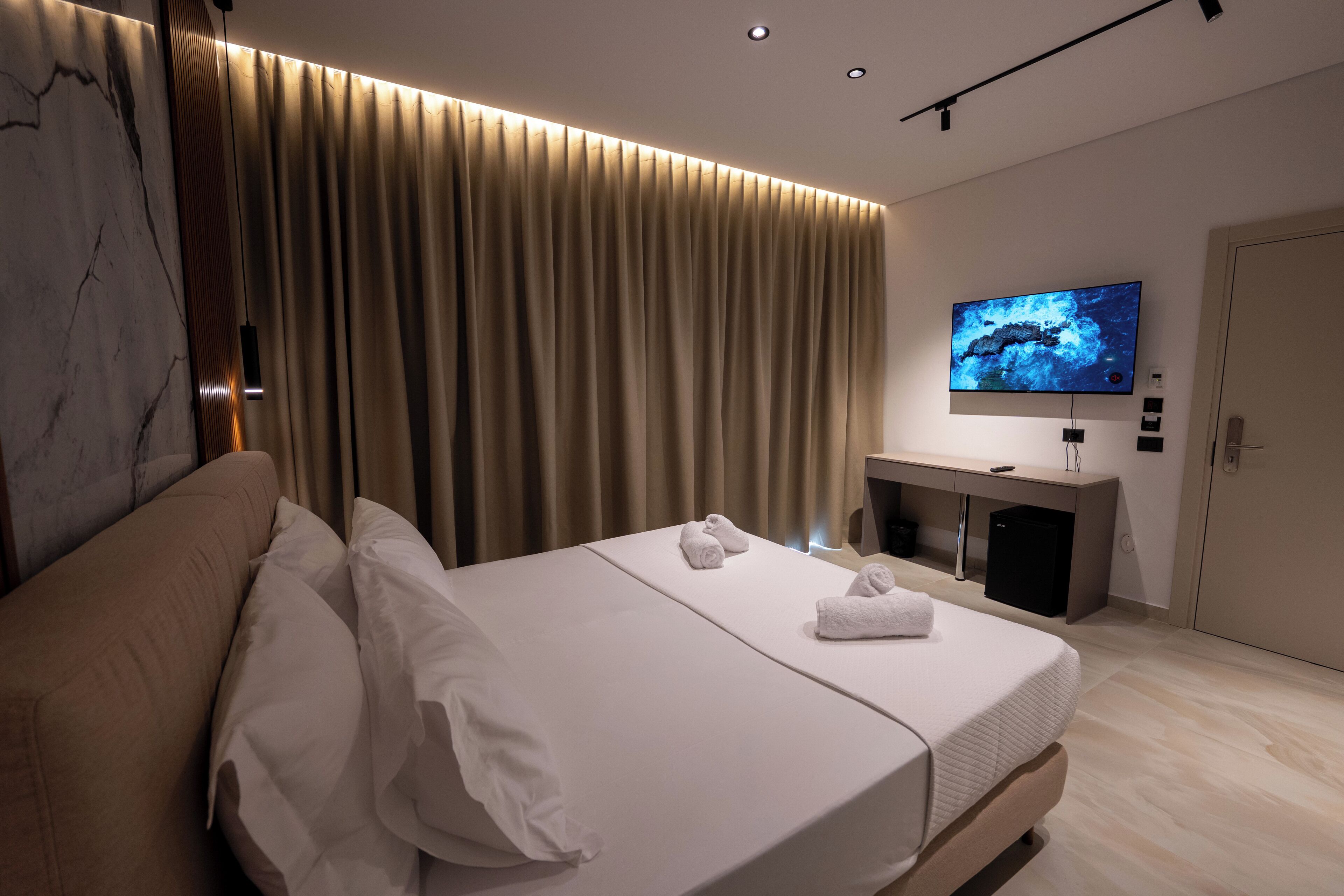 A modern hotel room with a large bed, a TV mounted on the wall, and a desk with a computer.