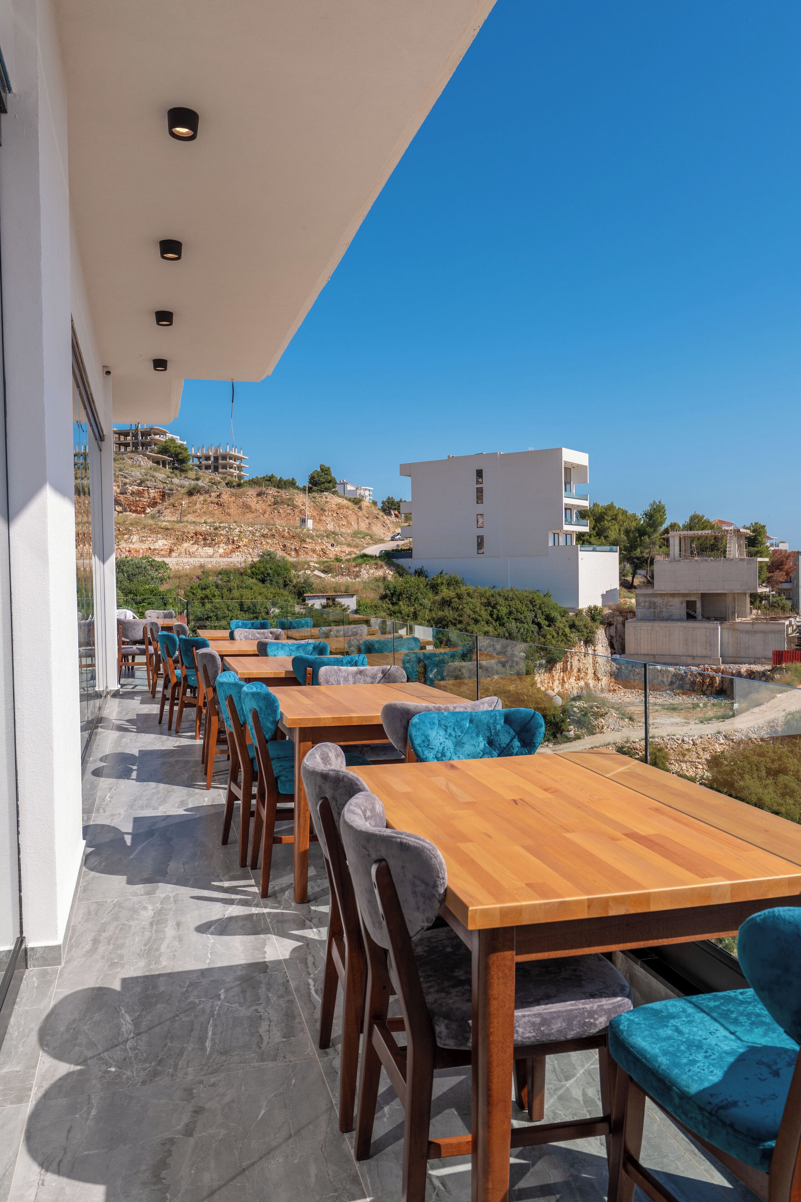 An outdoor dining area with wooden tables and chairs, a clear view of the surrounding landscape, and a modern building in the background.
