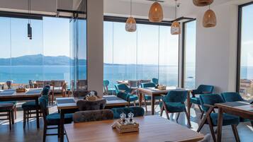 A modern dining area with tables and chairs, large windows with a view of the sea, and hanging pendant lights.