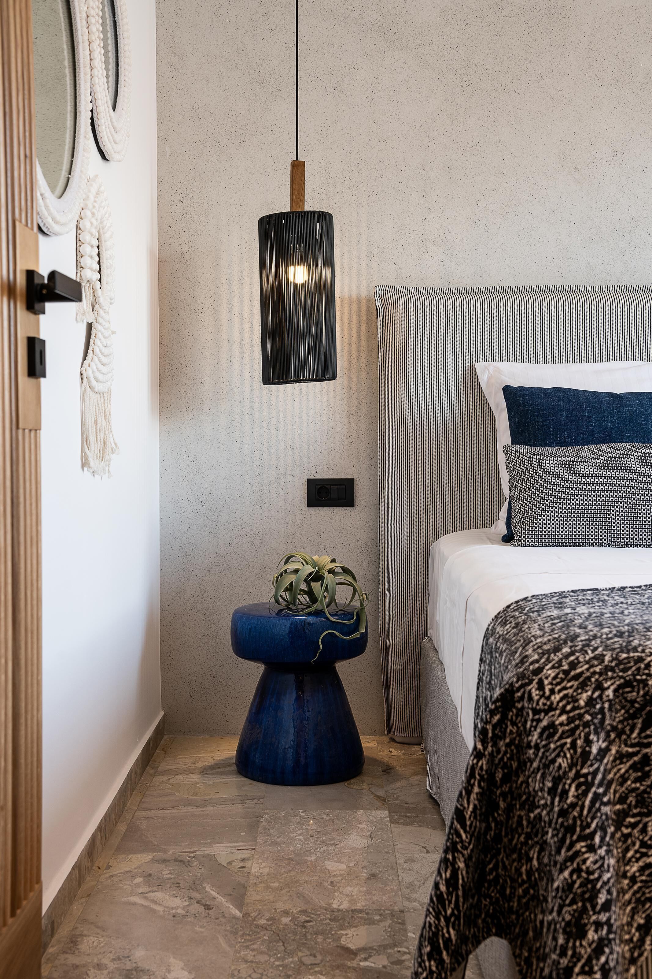 A modern bedroom with a bed, a hanging light fixture, a blue stool, and a macrame wall hanging.