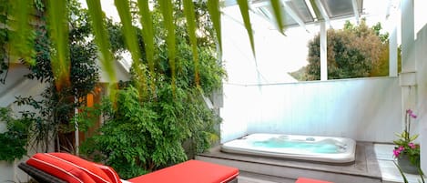 Un espacio al aire libre iluminado por el sol con dos sillones rojos, un jacuzzi y abundante vegetación.
