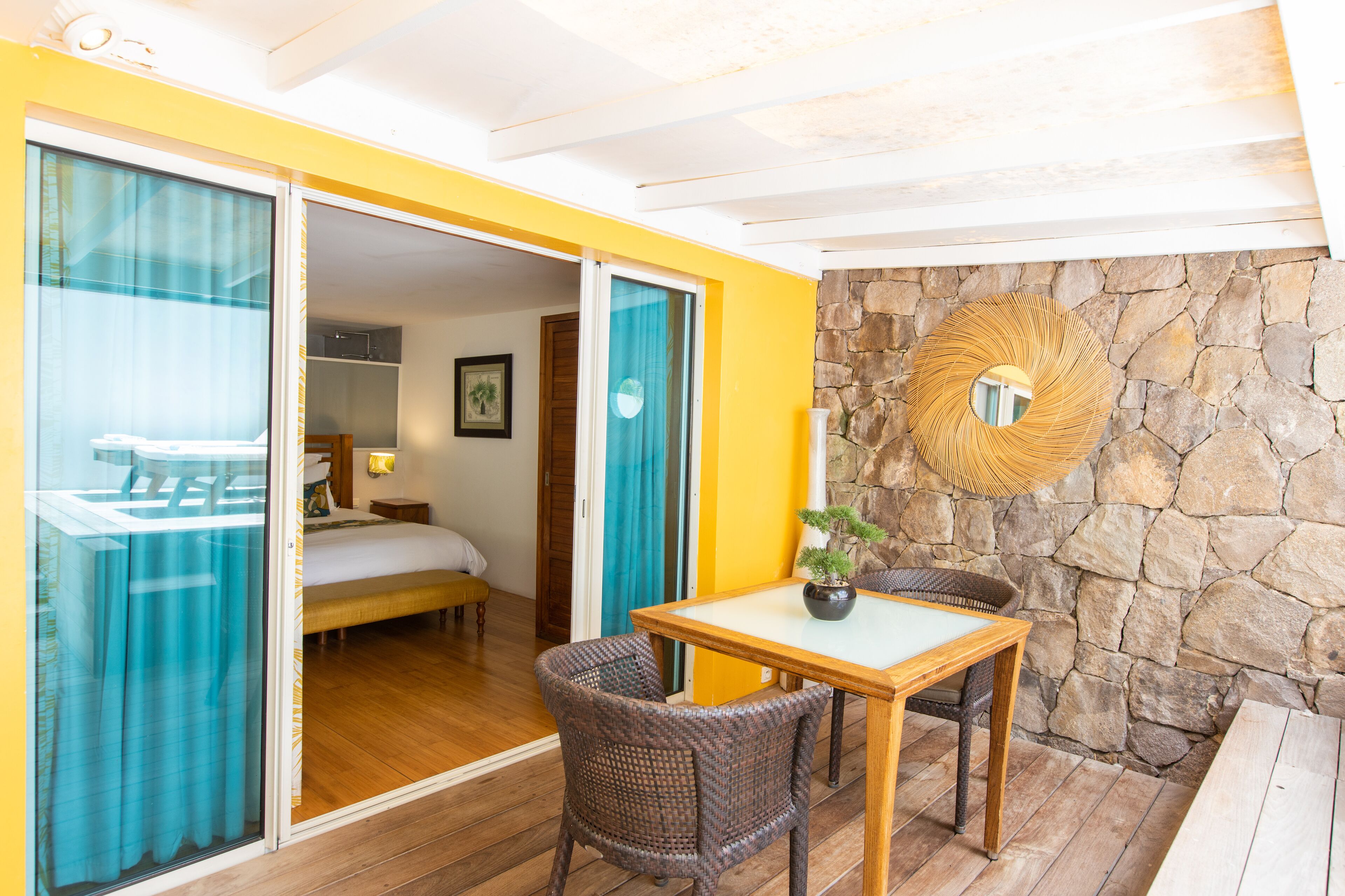 A room with a bed, a table with chairs, a potted plant, and a stone wall with a circular mirror.