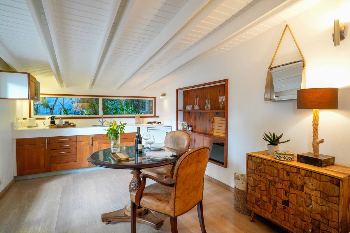 Een moderne keuken met houten kasten, een eettafel met stoelen, een groot raam met uitzicht op groen, en een houten kast met decoratieve items.
