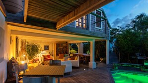 A wooden deck with outdoor seating, a pool with green lighting, and a building with large windows and a balcony. - La Maison Fleur de Lune (St. Barthelemy)