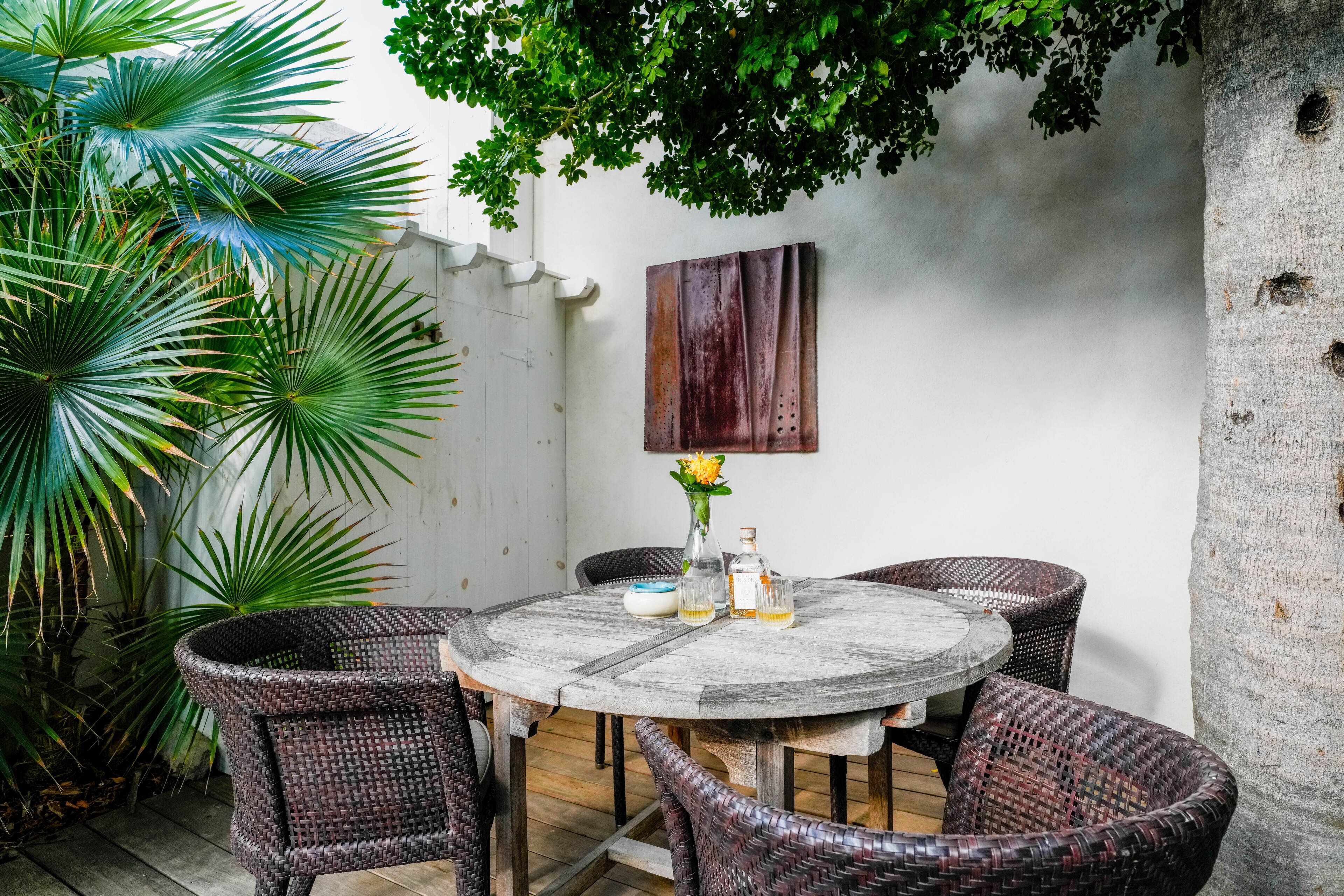 A round table with wicker chairs, a vase with flowers, and two bottles on the table.