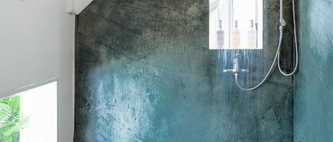 A shower with a handheld showerhead and a handheld hose, a window with a view of greenery, and a wooden floor.