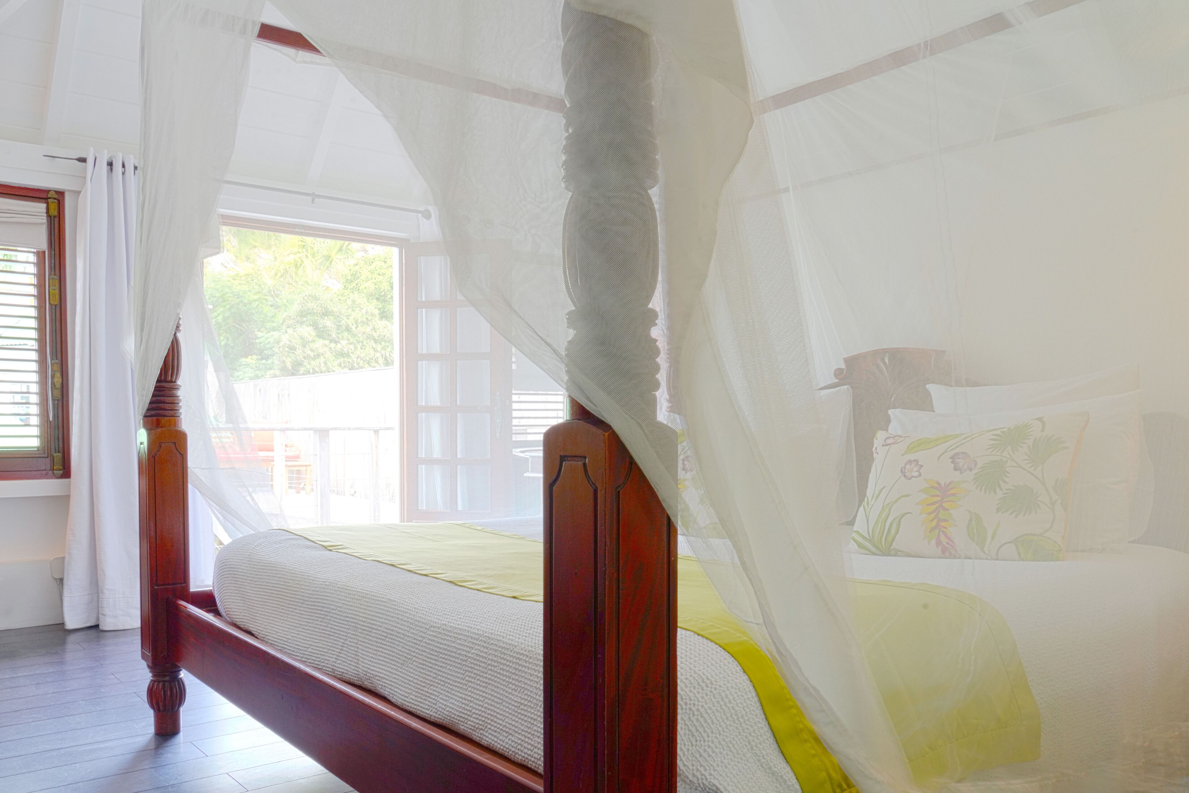 A bed with a wooden frame and a canopy, a window with curtains, and a view of greenery outside.